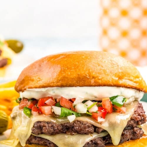 A cheeseburger with two beef patties, melted cheese, pico de gallo, and creamy sauce on a toasted bun, served on a white plate with crinkle-cut fries in the background.