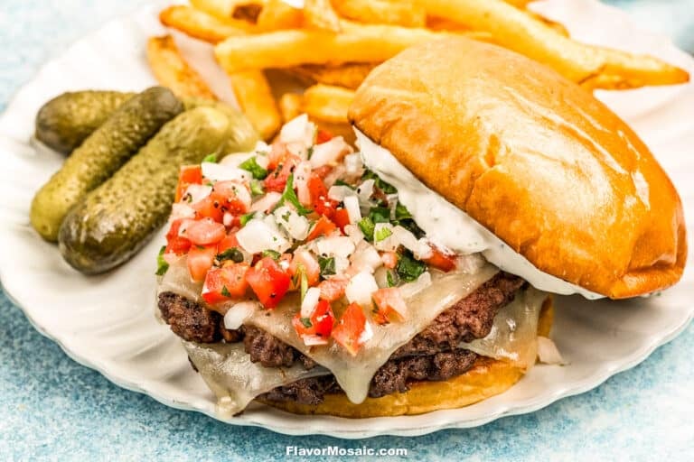 A cheeseburger topped with diced tomatoes, onions, and herbs sits on a plate with pickles, French fries, and a brioche bun spread with white sauce.