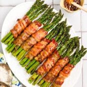 A white platter displays bacon-wrapped asparagus bundles drizzled with honey glaze. A small bowl of honey with a dipper and two vintage serving spoons rest nearby on the tiled surface.