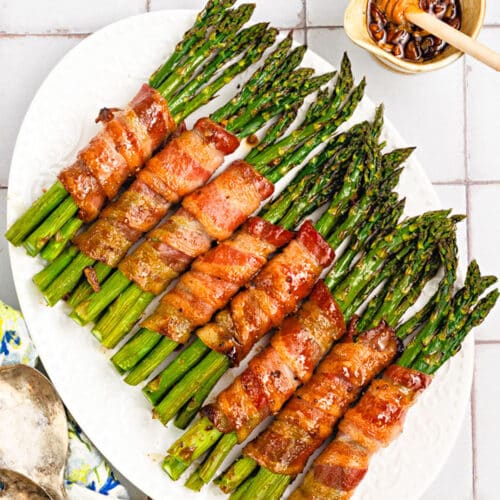 A white platter displays bacon-wrapped asparagus bundles drizzled with honey glaze. A small bowl of honey with a dipper and two vintage serving spoons rest nearby on the tiled surface.