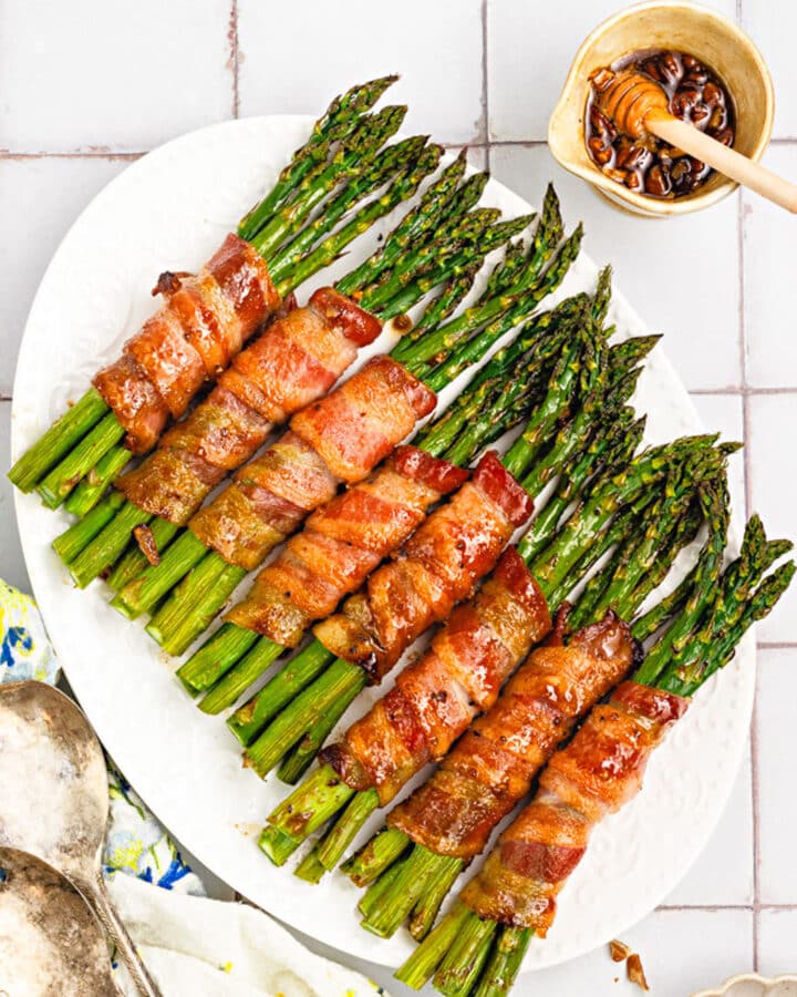 A white platter displays bacon-wrapped asparagus bundles drizzled with honey glaze. A small bowl of honey with a dipper and two vintage serving spoons rest nearby on the tiled surface.