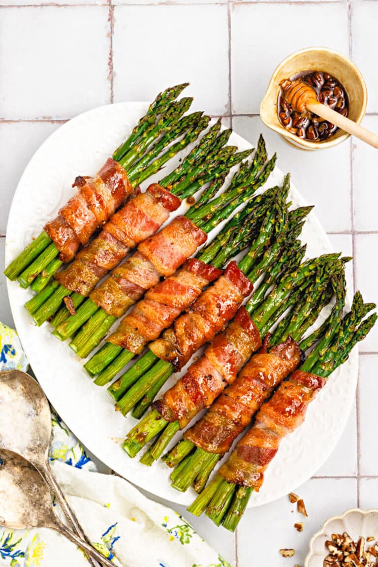 A white platter displays bacon-wrapped asparagus bundles drizzled with honey glaze. A small bowl of honey with a dipper and two vintage serving spoons rest nearby on the tiled surface.