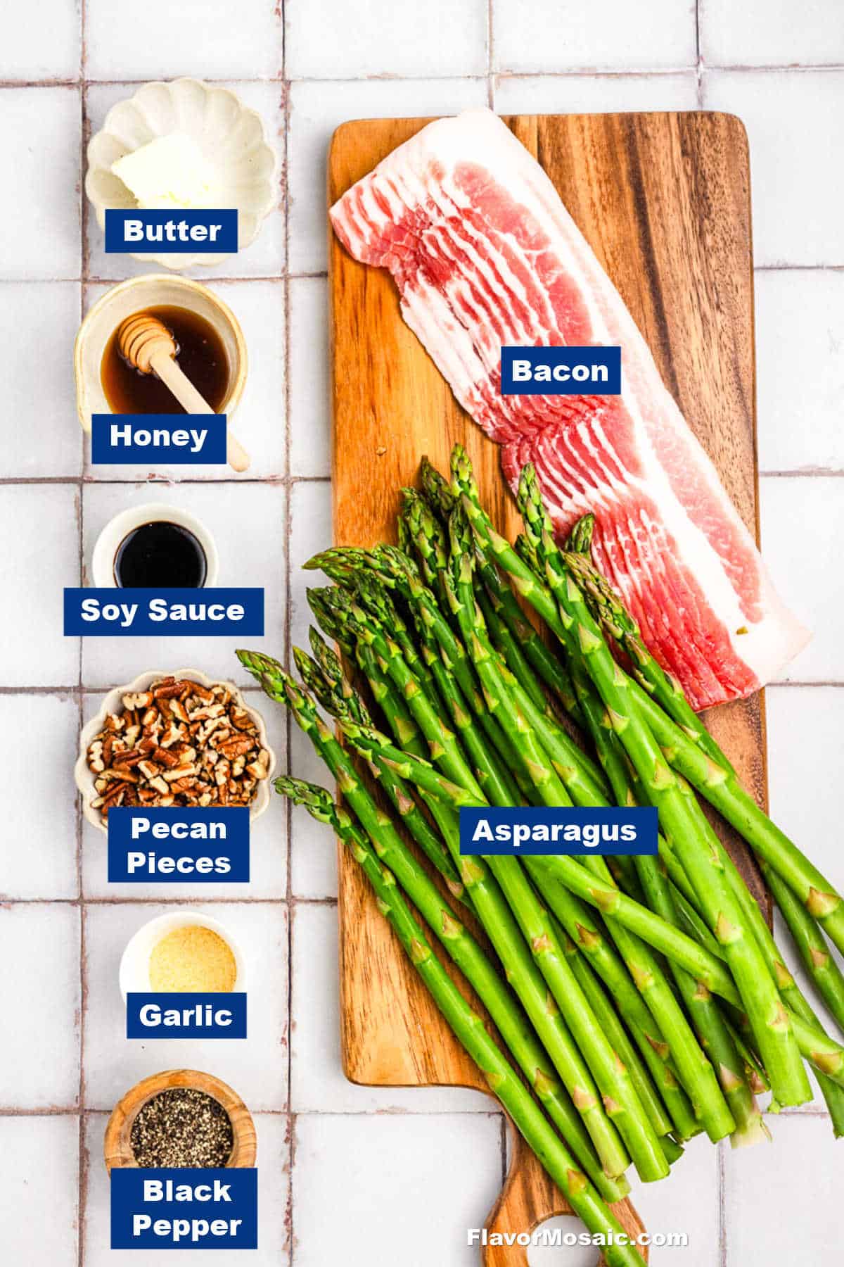 Ingredients for Bacon-Wrapped Asparagus arranged on a white tile surface: uncooked bacon, asparagus spears, butter, honey, soy sauce, pecan pieces, chopped garlic, and ground black pepper—each labeled with blue tags.