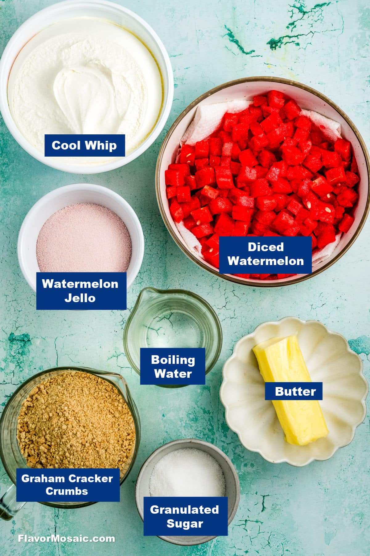 Ingredients for a dessert are arranged on a light blue surface: Cool Whip, diced watermelon, watermelon Jello, boiling water, butter, graham cracker crumbs, and granulated sugar, each labeled.