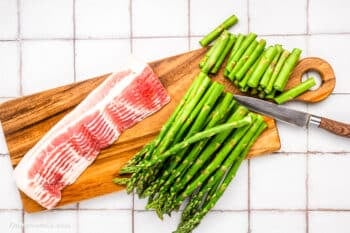 Bacon-wrapped asparagus bundles and chopped spears sit on a wooden cutting board with a knife, ready for prepping on a white tiled surface.