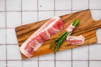 Three raw bacon strips sit on a wooden cutting board alongside several bacon-wrapped asparagus bundles. The neatly wrapped Bacon-Wrapped Asparagus rests on the board, which is placed atop a white tiled surface.