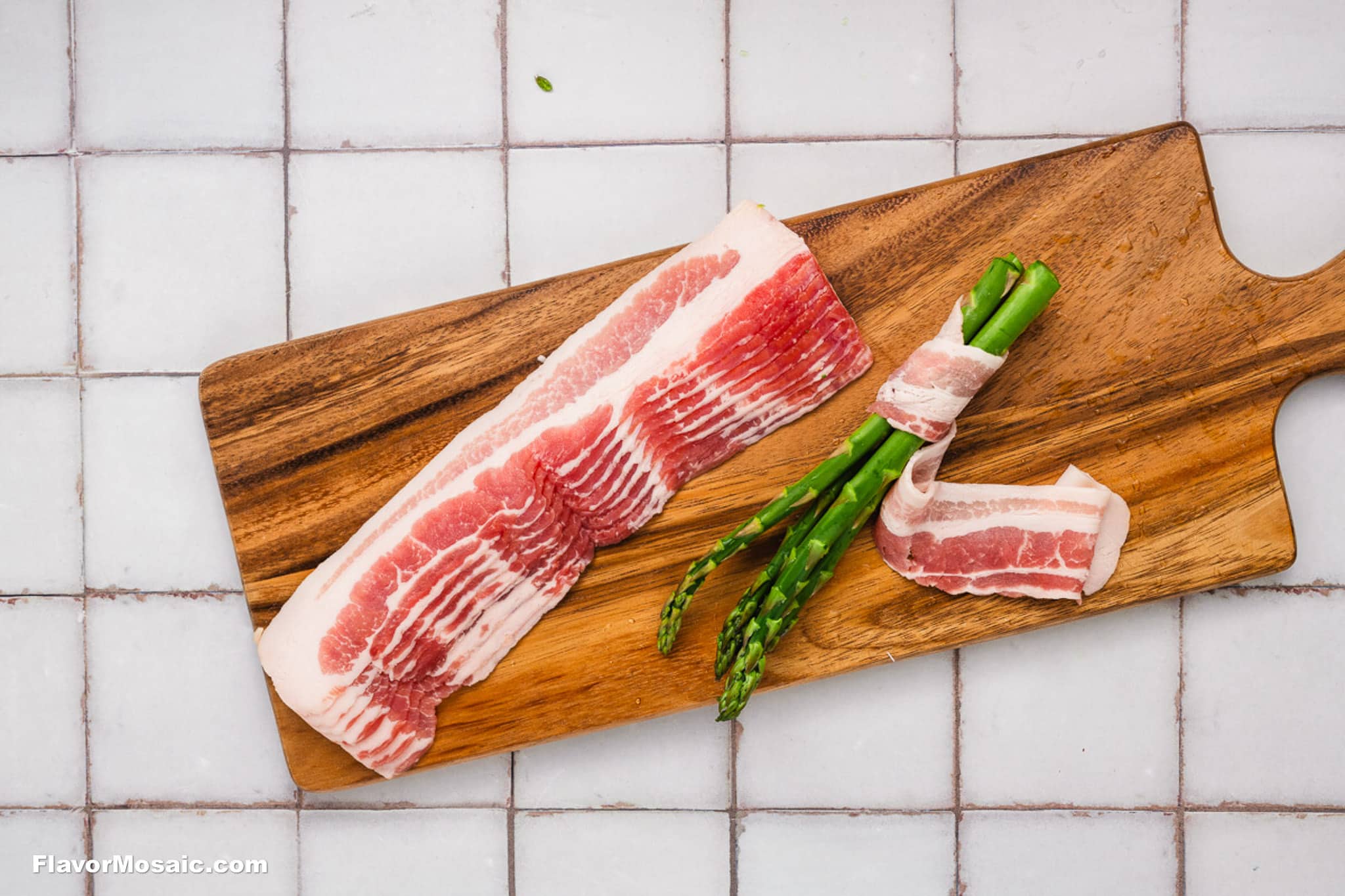 Three raw bacon strips sit on a wooden cutting board alongside several bacon-wrapped asparagus bundles. The neatly wrapped Bacon-Wrapped Asparagus rests on the board, which is placed atop a white tiled surface.