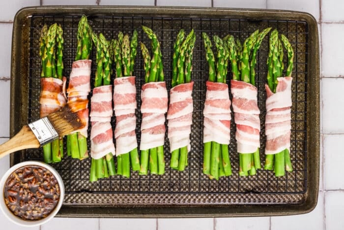 Bacon-wrapped asparagus bundles are arranged on a wire rack over a baking pan. A hand uses a brush to apply sauce from a small bowl to the Bacon-Wrapped Asparagus before baking.