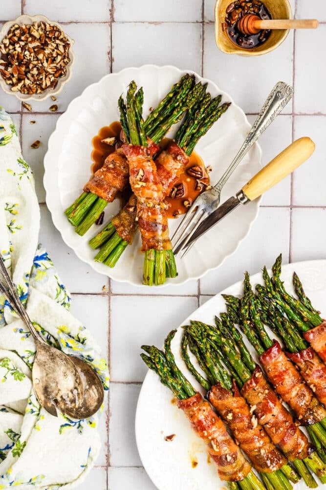 Bacon-wrapped asparagus bundles are served on white plates, drizzled with sauce and sprinkled with chopped nuts, accompanied by a fork, spoon, floral napkin, and a small bowl of nuts on a tiled surface.