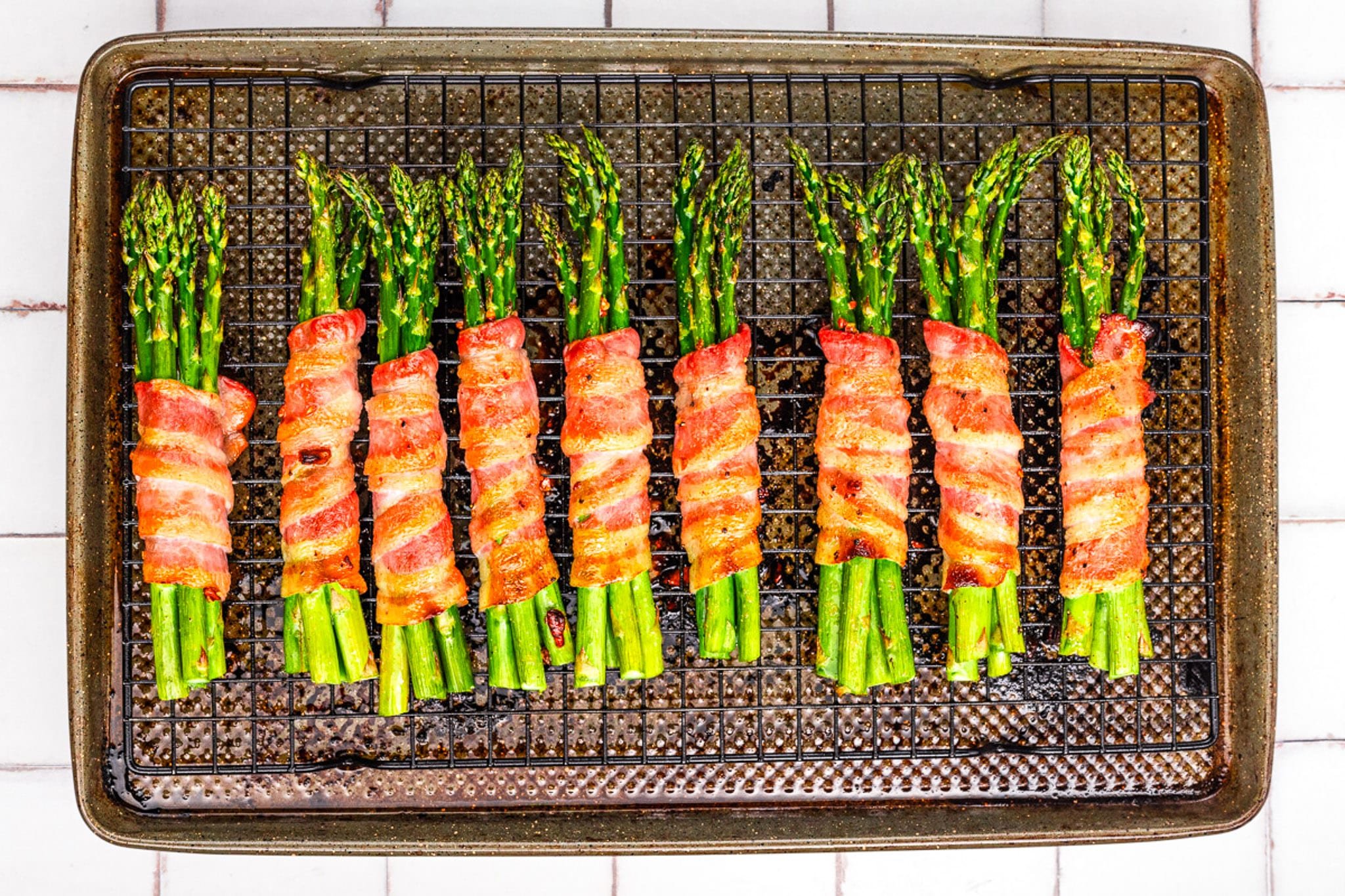 Freshly cooked bacon-wrapped asparagus bundles are arranged in neat rows on a wire rack over a baking sheet, showcased on a tiled surface.
