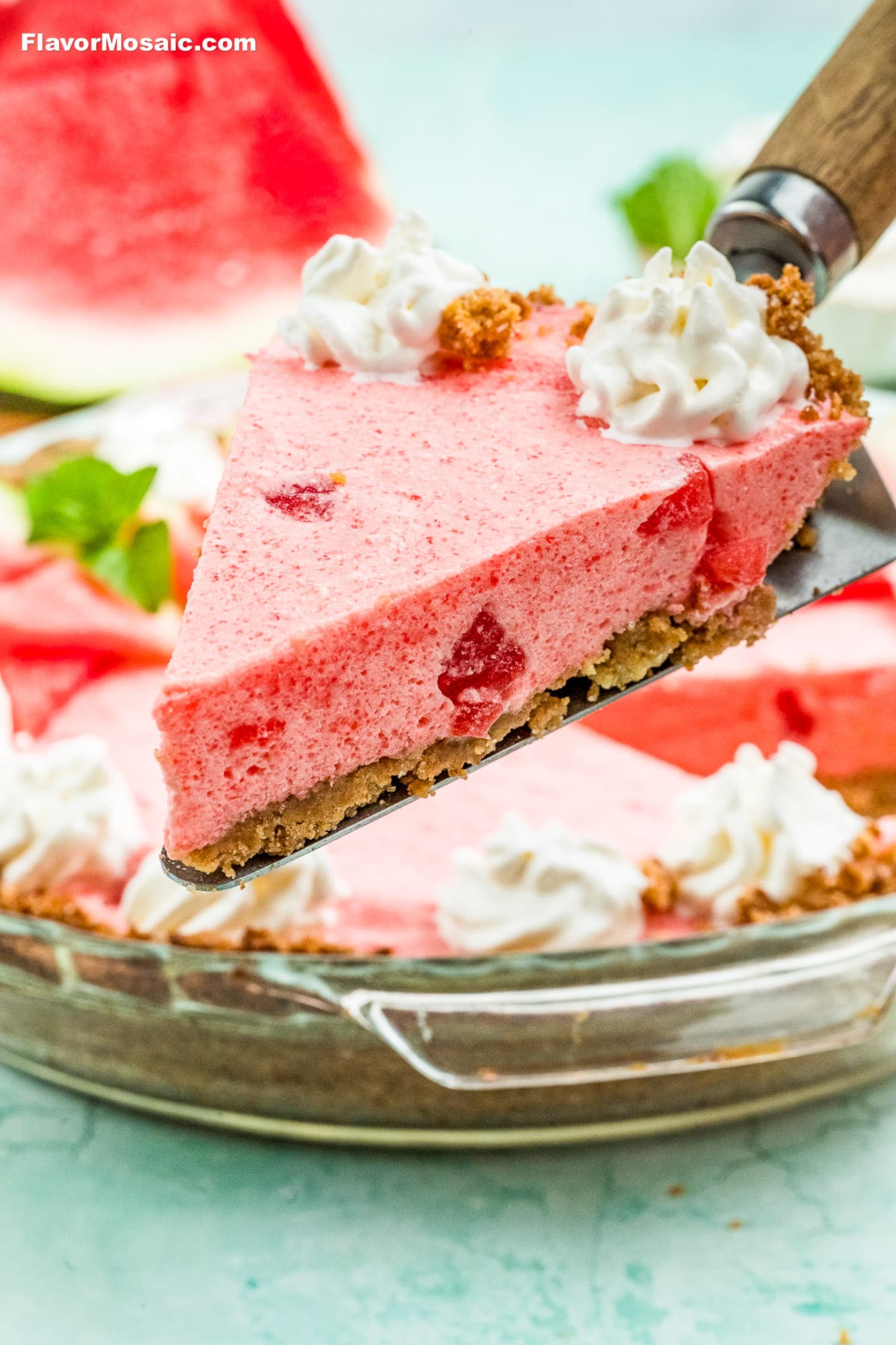A close-up of a slice of pink watermelon pie with whipped cream and graham cracker crust, being lifted from a glass pie dish. There are pieces of watermelon in the pie and a large watermelon wedge in the background.