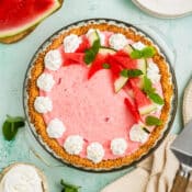 A pink watermelon pie with a graham cracker crust, decorated with whipped cream, fresh watermelon slices, and mint leaves, sits on a light green surface with plates, forks, a pie server, and more watermelon nearby.