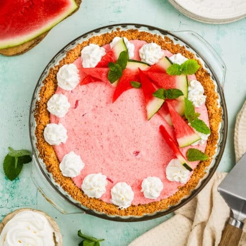 A pink watermelon pie with a graham cracker crust, decorated with whipped cream, fresh watermelon slices, and mint leaves, sits on a light green surface with plates, forks, a pie server, and more watermelon nearby.