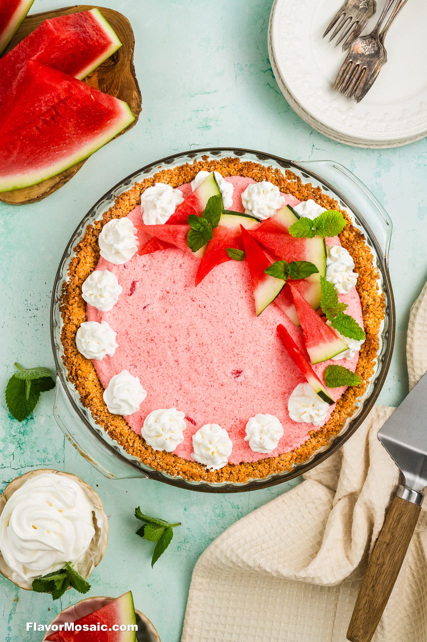 A pink watermelon pie with a graham cracker crust, decorated with whipped cream, fresh watermelon slices, and mint leaves, sits on a light green surface with plates, forks, a pie server, and more watermelon nearby.