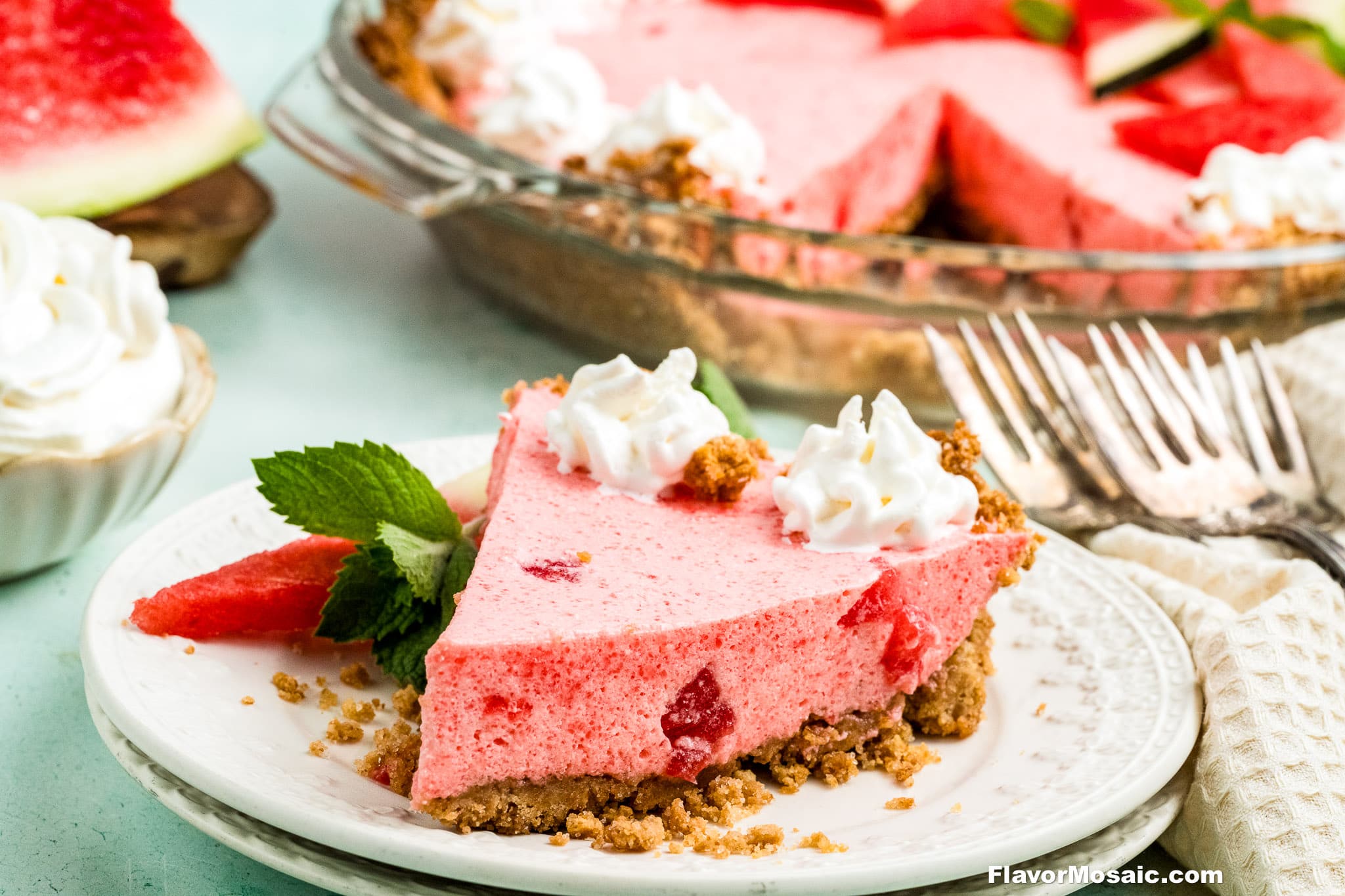 A slice of pink watermelon pie with graham cracker crust, topped with whipped cream and mint, sits on a white plate with a fork. The rest of the pie and a bowl of whipped cream are in the background.