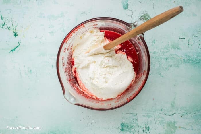 A glass measuring cup filled with a layer of whipped cream or white mixture atop a red liquid, with a wooden spoon resting inside, sits on a light turquoise, textured surface.