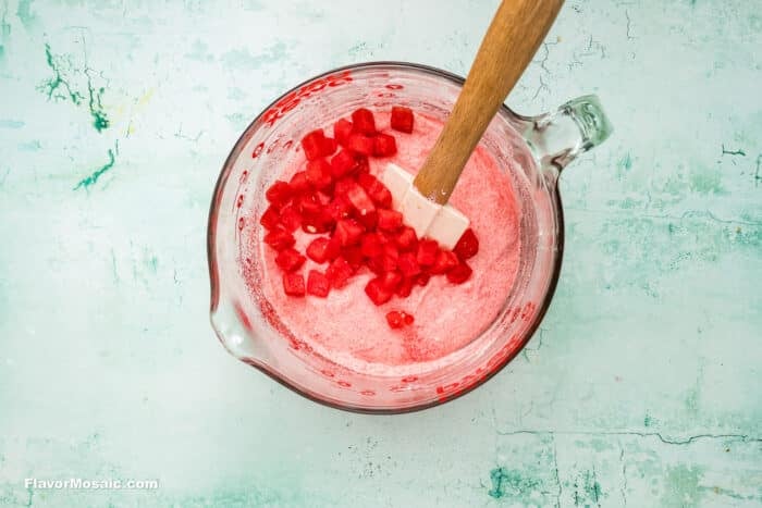 A glass mixing bowl with a pink foamy mixture and diced watermelon, stirred with a white spatula, sits on a light blue textured surface.