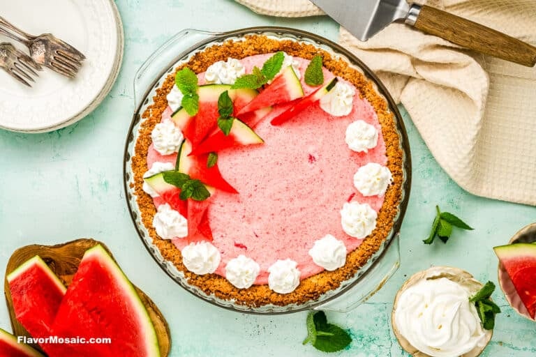 A watermelon pie with a pink filling in a graham cracker crust, topped with whipped cream dollops, watermelon slices, and mint leaves, sits on a light blue surface with plates, utensils, and extra watermelon nearby.