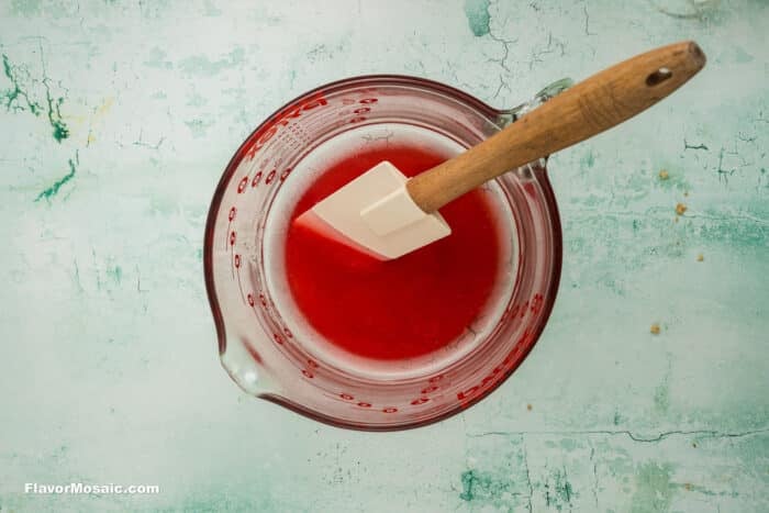 A glass measuring cup filled with red liquid and a white spatula with a wooden handle resting inside, set on a light textured surface.