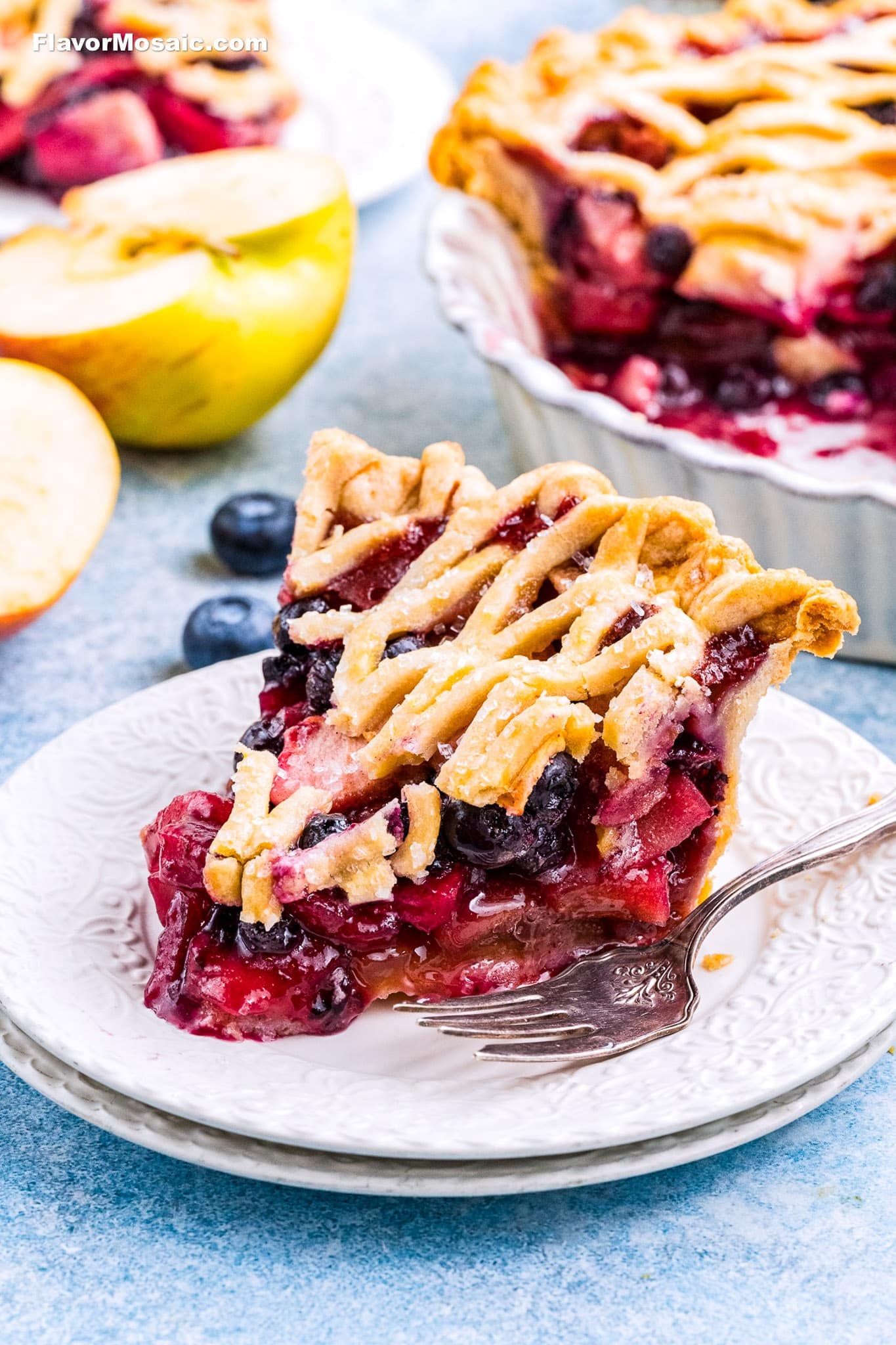 A slice of berry and apple pie with a lattice crust sits on a white plate with a fork. The pie is filled with mixed berries and apples, and there are fresh apple slices and more pie in the background.