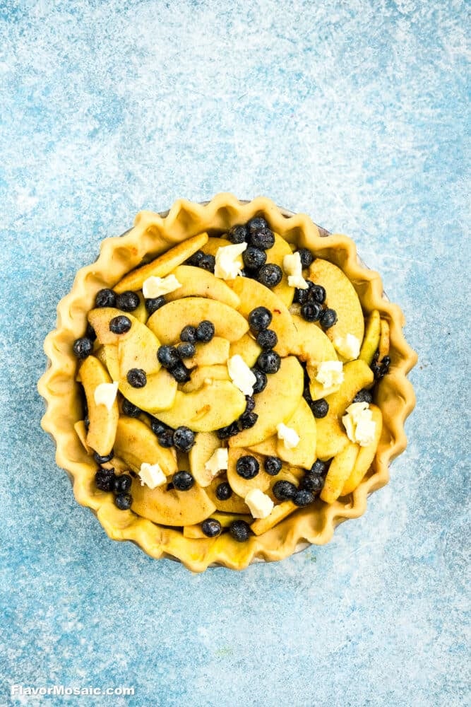 Unbaked pie with a fluted crust, filled with sliced apples, blueberries, and small cubes of butter, on a blue textured background.
