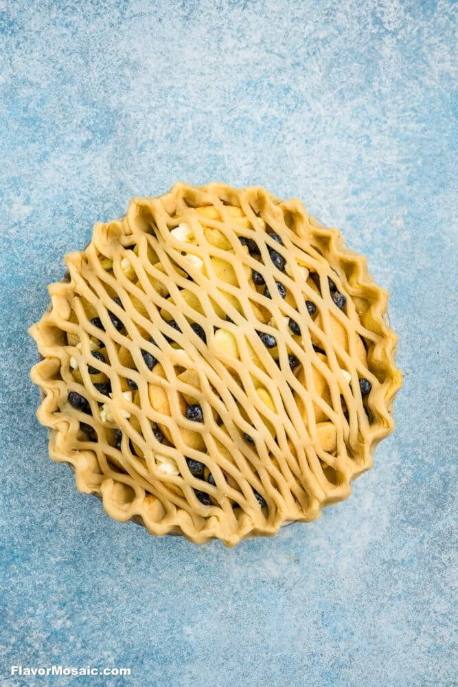 Unbaked lattice-top pie filled with blueberries and apple chunks on a blue textured surface. The pie has a crimped edge and the lattice crust is woven diagonally across the top.