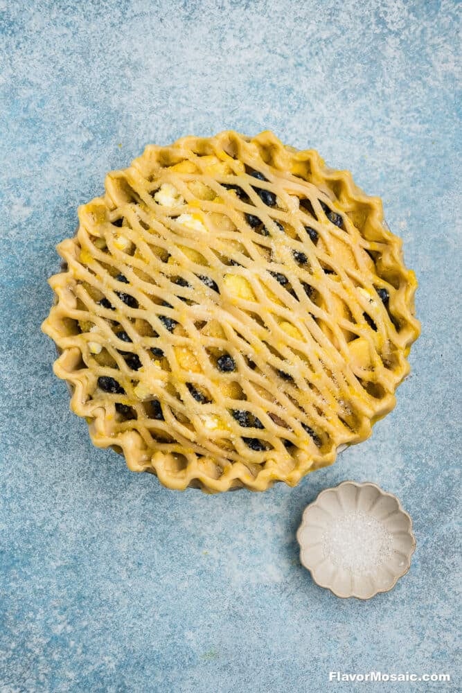 A lattice-topped unbaked pie filled with fruit sits on a blue textured surface, next to a small scalloped dish containing coarse sugar.