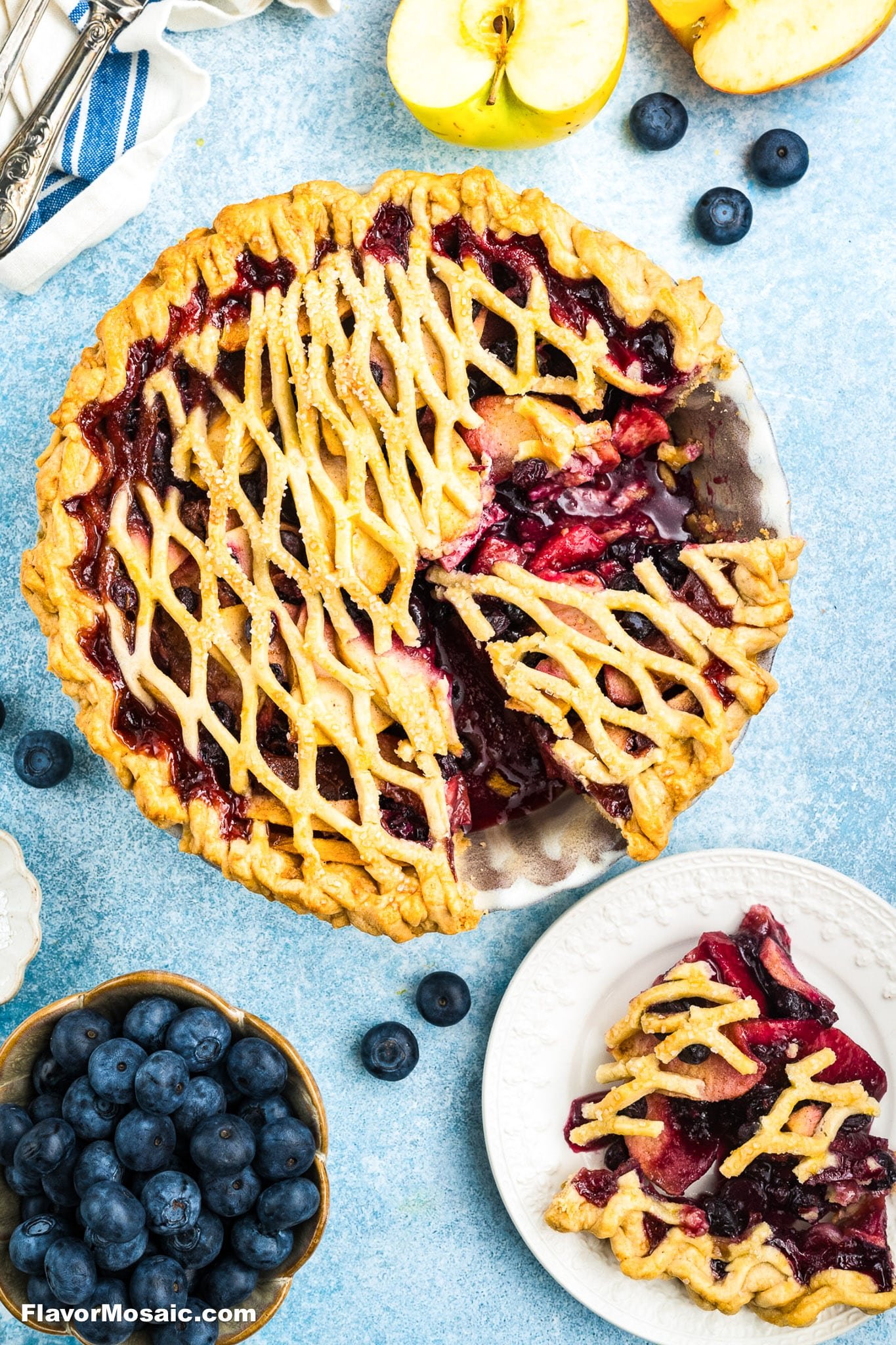 A lattice-topped mixed berry pie with a slice served on a white plate, surrounded by fresh blueberries and sliced apples on a light blue surface.