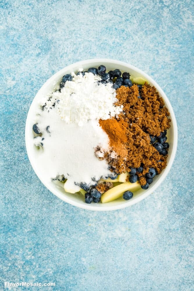 A white bowl filled with blueberries, sliced apples, granulated sugar, brown sugar, cinnamon, and powdered sugar, all separated in sections, set on a blue textured surface.