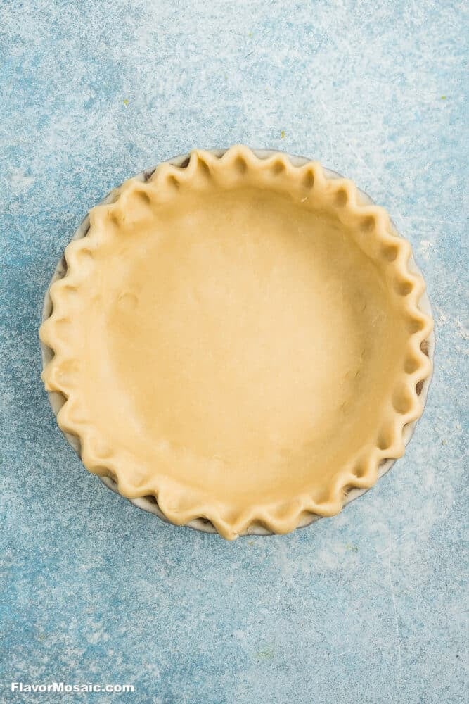 Unbaked Apple Blueberry Pie crust with fluted edges in a ceramic pie dish, placed on a light blue textured surface.