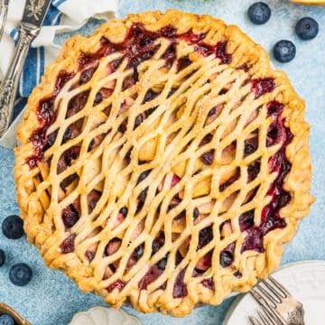 A freshly baked pie with a golden lattice crust, filled with blueberries and sliced apples, sits on a blue surface surrounded by fresh blueberries, apple slices, plates, forks, and a small bowl of sugar.