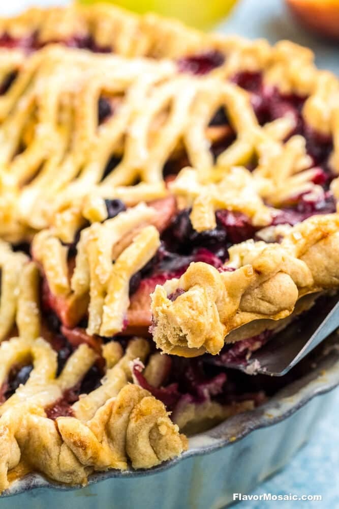 A close-up of a freshly baked Apple Blueberry Pie with a golden, crisscross lattice crust. A slice is being lifted out, revealing juicy berries and apples nestled inside the flaky pastry.
