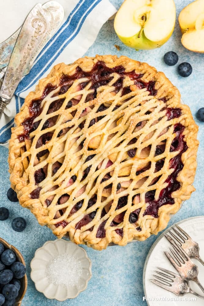 A freshly baked blueberry and apple pie with a golden lattice crust sits on a blue surface, surrounded by apple slices, fresh blueberries, a small bowl of sugar, forks, and vintage knives.