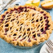A freshly baked pie with a golden, lattice crust is displayed on a blue surface, surrounded by blueberries, sliced apples, a plate, and forks. The pie filling appears to be a mix of berries and apples.