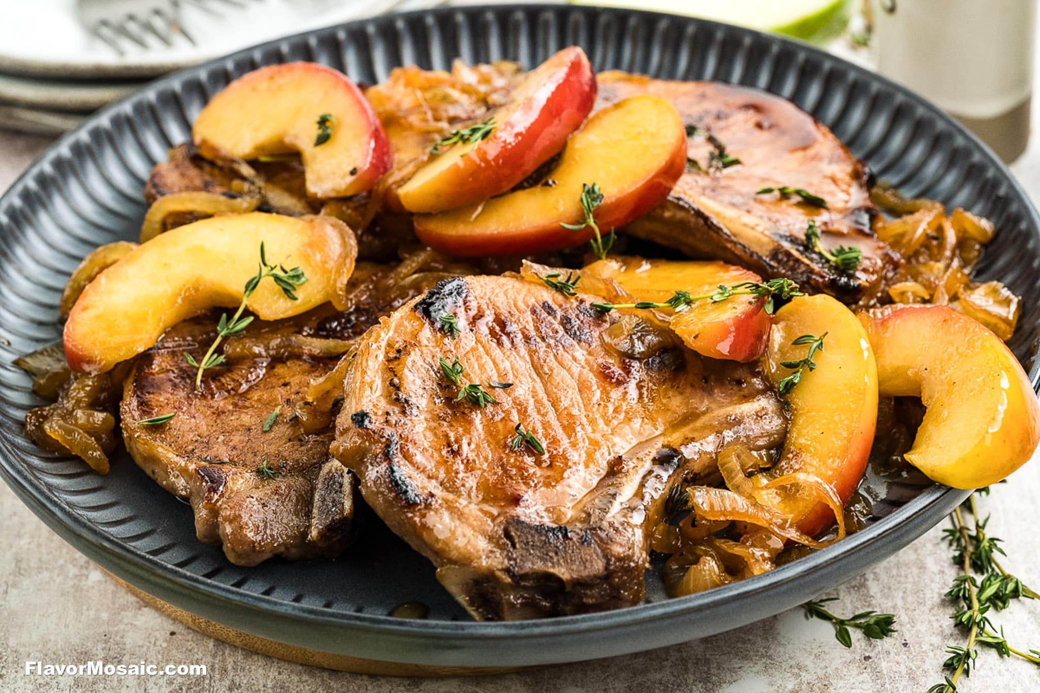 A black plate with grilled pork chops topped with caramelized onions, sliced peaches, and fresh thyme sprigs, served on a light-colored tabletop.