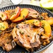 A plate of cooked pork chops garnished with sliced apples, caramelized onions, and fresh thyme, served on a dark dish with lemon wedges and salt and pepper shakers in the background.