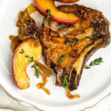 A cooked pork chop garnished with caramelized onions, fresh thyme, and sliced red apples is served on a white plate.