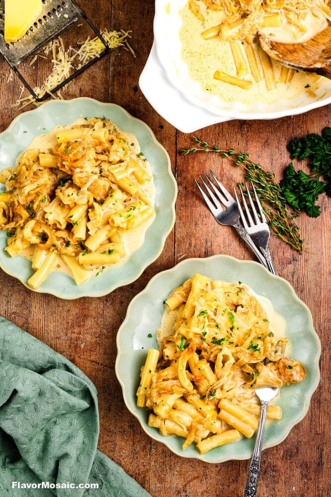 Two green plates filled with creamy pasta and chicken sit on a wooden table, surrounded by herbs, shredded cheese, a serving dish, and two forks. A green napkin and scattered parsley add color to the scene.