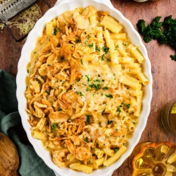 Baked macaroni and cheese in a white oval dish, topped with crispy fried onions and chopped parsley, placed on a wooden table with forks, a napkin, cheese, and herbs nearby.