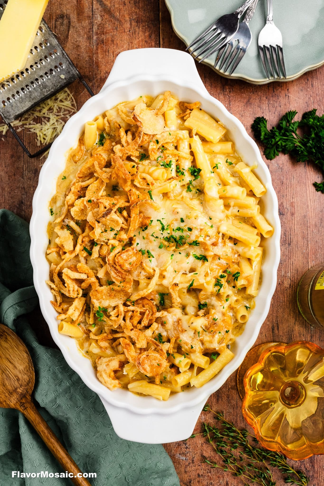 Baked macaroni and cheese in a white oval dish, topped with crispy fried onions and chopped parsley, placed on a wooden table with forks, a napkin, cheese, and herbs nearby.