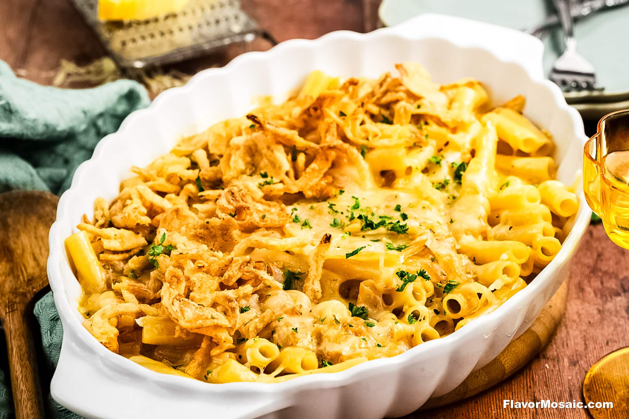 A white casserole dish filled with baked pasta topped with melted cheese, crispy fried onions, and chopped parsley, sitting on a wooden table with utensils and a glass of drink nearby.