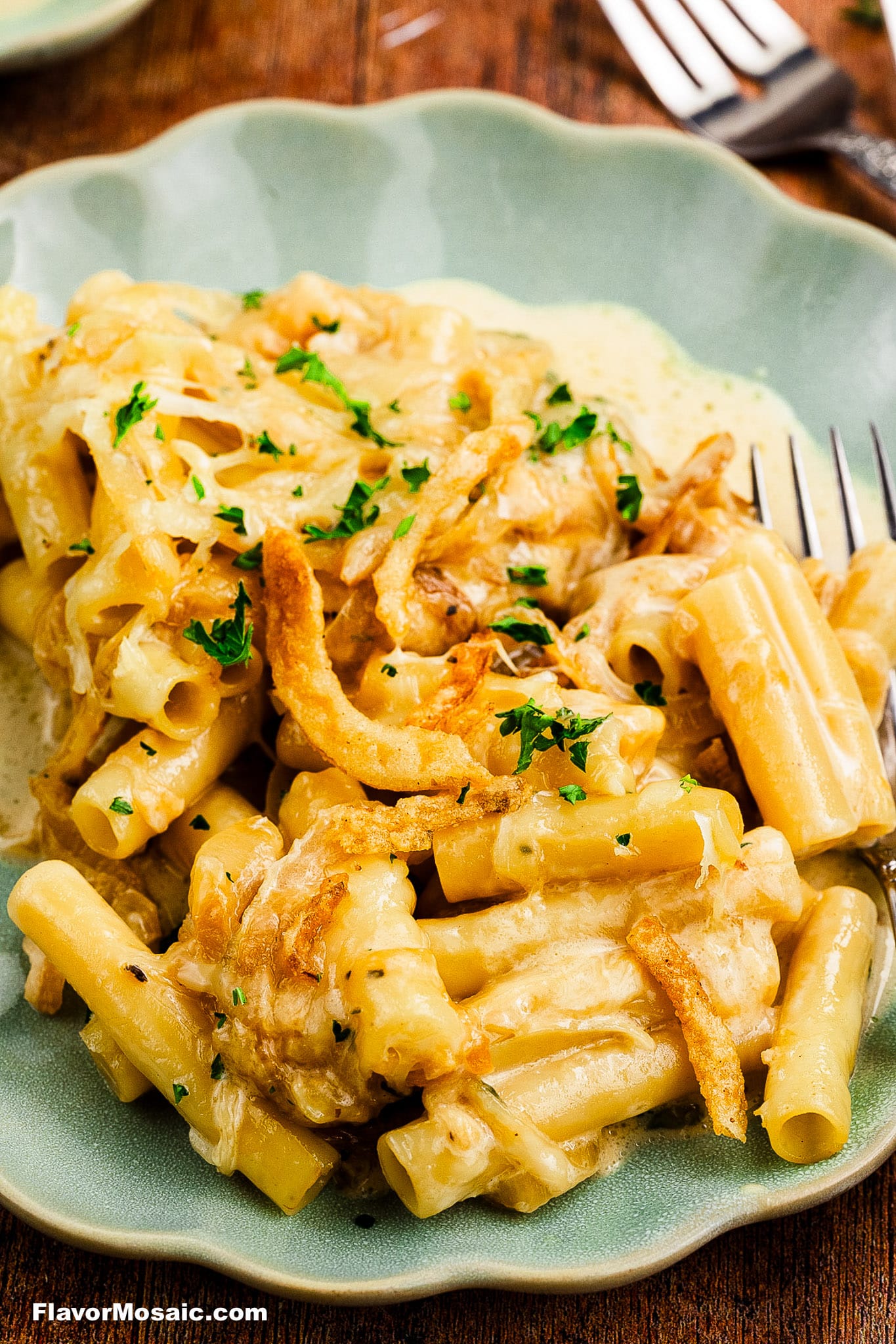 A close-up of creamy baked macaroni and cheese topped with crispy fried onions and chopped parsley, served on a green plate with a fork nearby.