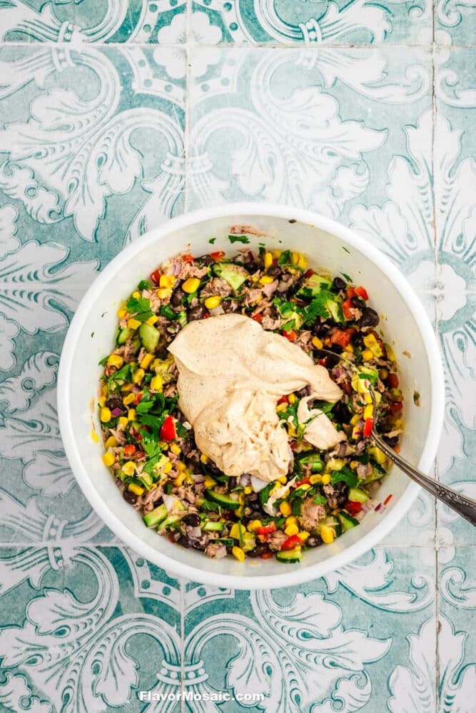 A white bowl filled with Mexican Tuna Salad-a colorful mixture of chopped vegetables, beans, and corn-topped with a dollop of creamy dressing. A spoon rests in the bowl on a decorative, teal patterned surface.
