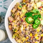 A bowl of creamy Mexican Tuna Salad with black beans, corn, avocado, red onion, and cilantro, garnished with jalapeño slices and extra cilantro, sits on a patterned surface.
