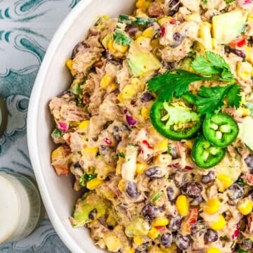 A bowl of creamy Mexican Tuna Salad with black beans, corn, avocado, red onion, and cilantro, garnished with jalapeño slices and extra cilantro, sits on a patterned surface.
