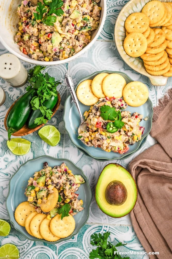 Colorful Mexican Tuna Salad with chopped veggies and herbs is served on blue plates with round crackers. Whole avocado, jalapeños, lime wedges, cilantro, and a bowl of salad are arranged around for a fresh, vibrant presentation.