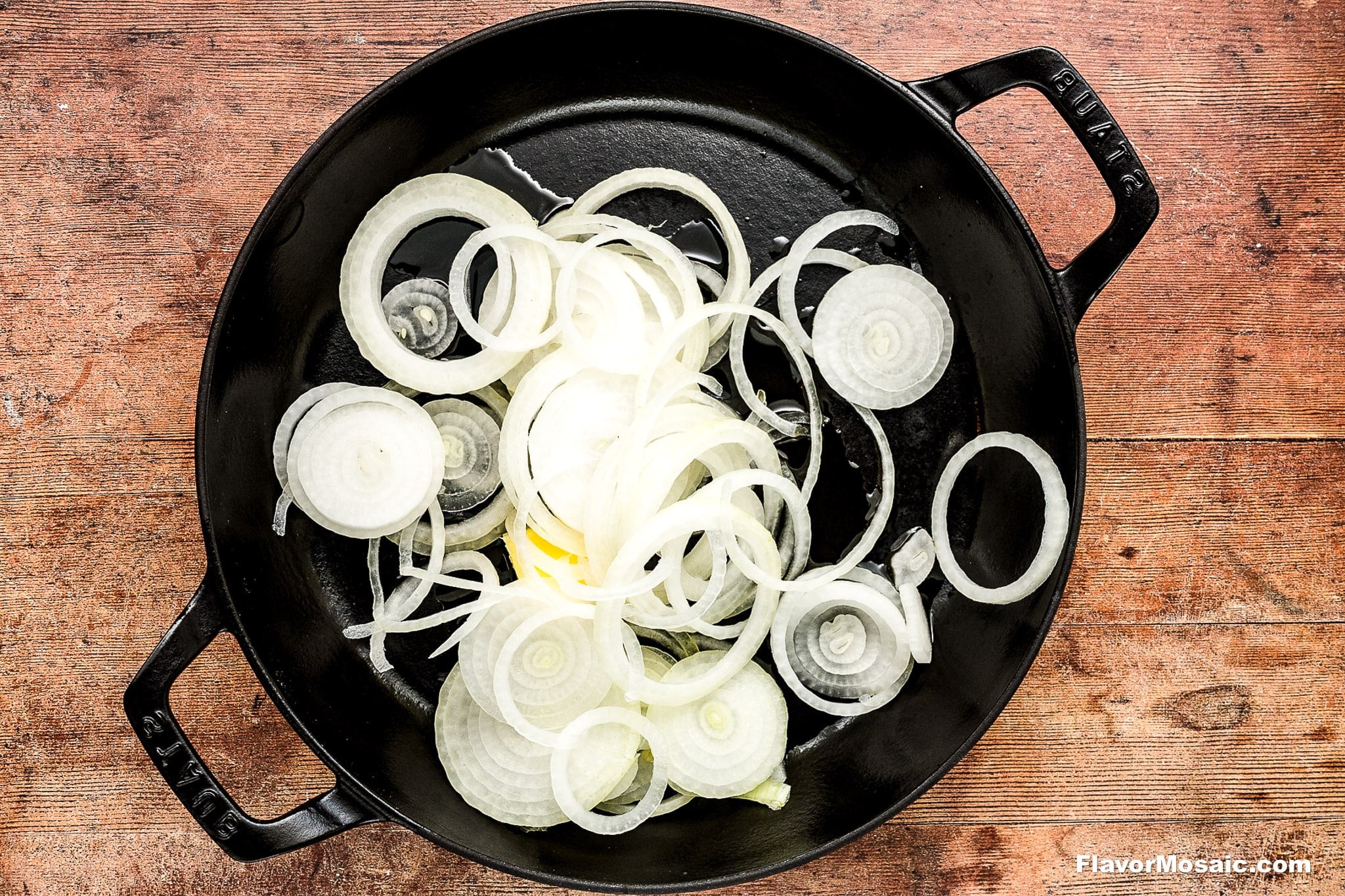 Thinly sliced white onions in a cast iron skillet with butter, and olive oil, ready to be cooked, placed on a rustic wooden surface.