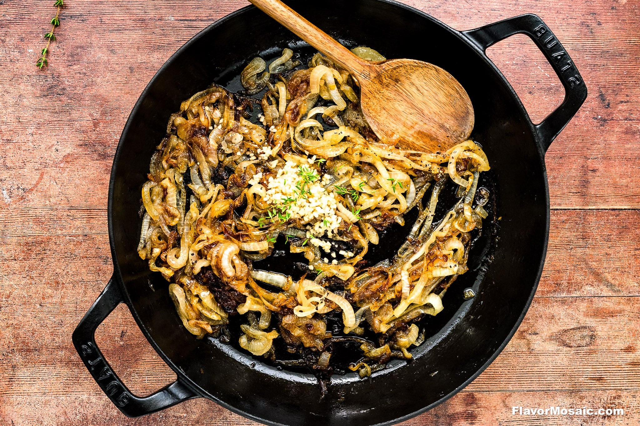 Sliced onions caramelizing in a black cast iron skillet with minced garlic and herbs in the center, and a wooden spoon resting on the side.