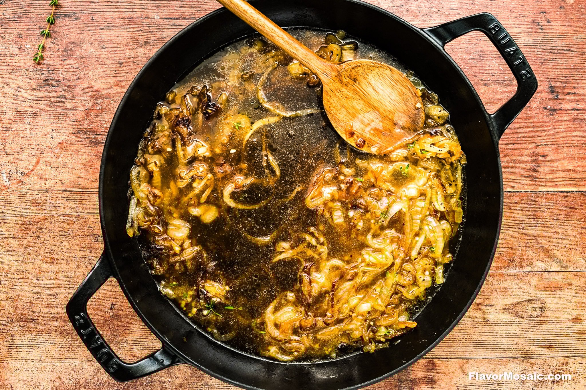 A black pot filled with caramelized onions simmering in broth, being stirred with a wooden spoon, sits on a wooden surface.