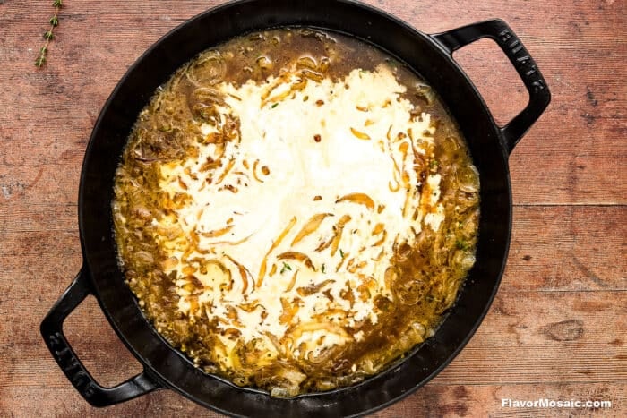 A black cast iron pot filled with French onion soup topped with melted cheese, sitting on a rustic wooden surface. The soup shows swirls of creamy cheese and visible cooked onions.